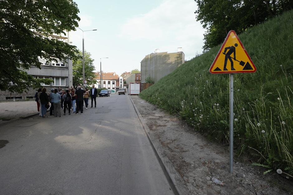 na zdjęciu fragment ulicy, po prawe widać żółty znak drogowy informujący o robotach ziemnych, w tle po lewej widać grupę kilkunastu osób które stoją w kółku i rozmawiają