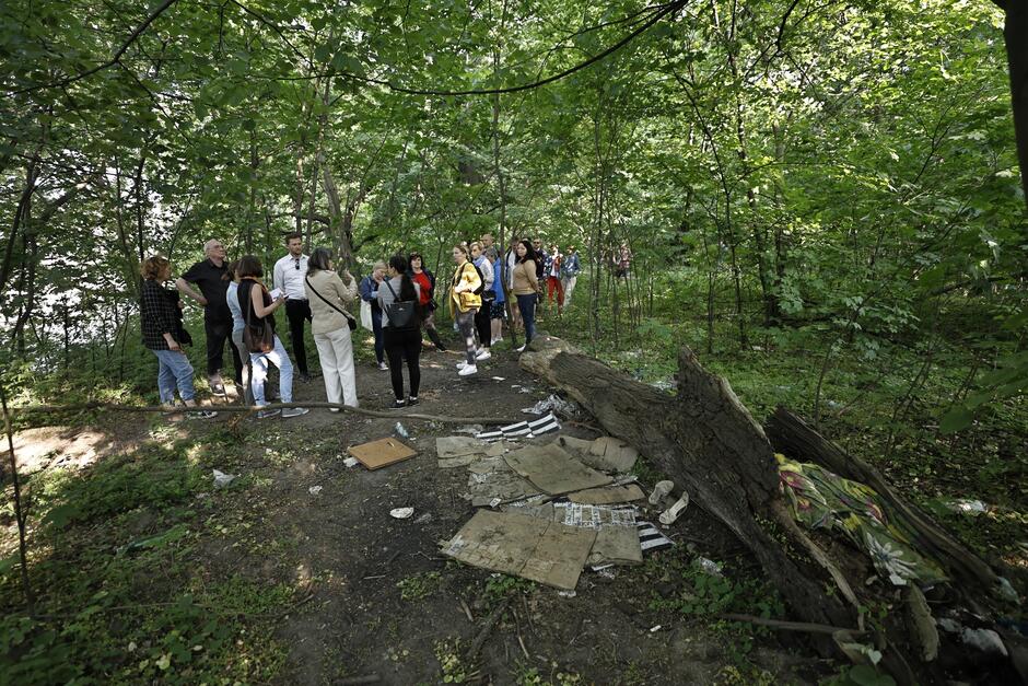na zdjęciu fragment lasu, widać trochę śmieci leżących na ziemi, w tle stoi grupka osób i rozmawia