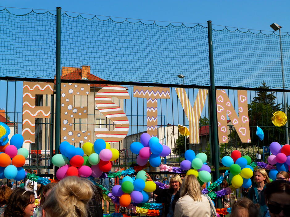 na zdjęciu kilkadziesiąt kolorowych balonów zawieszonych na siatce ochronnych przy boisku sportowym, przed nimi widać kilka stojących osób