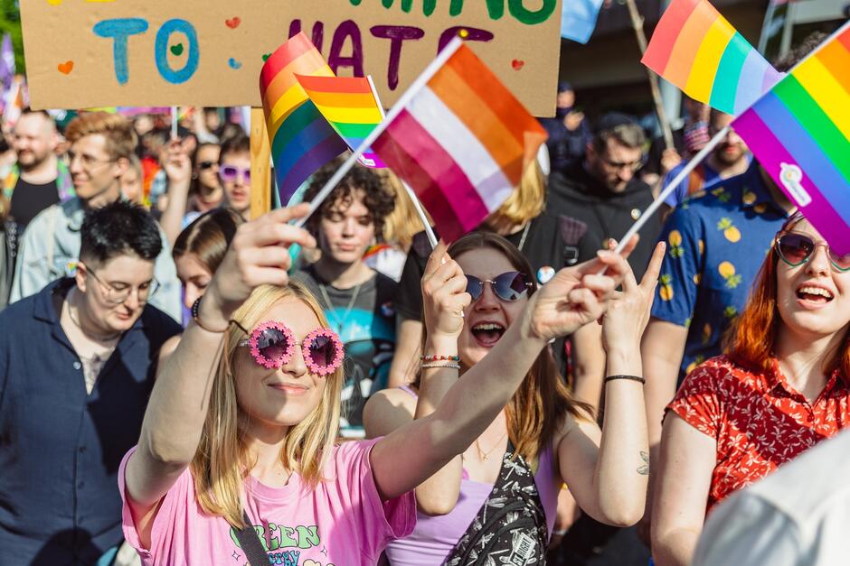 na zdjęciu kilka młodych dziewczyn na pierwszym planie idących w manifestacji, jedna z nich ma różowe okulary przeciwsłoneczne, trzyma kolorowe mini flagi w rękach