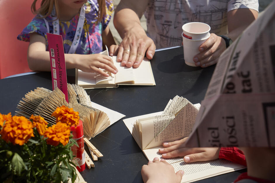 stół warsztatowy, mała dziewczynka z pomocą dorosłego składa kartki w notatniku lub książce