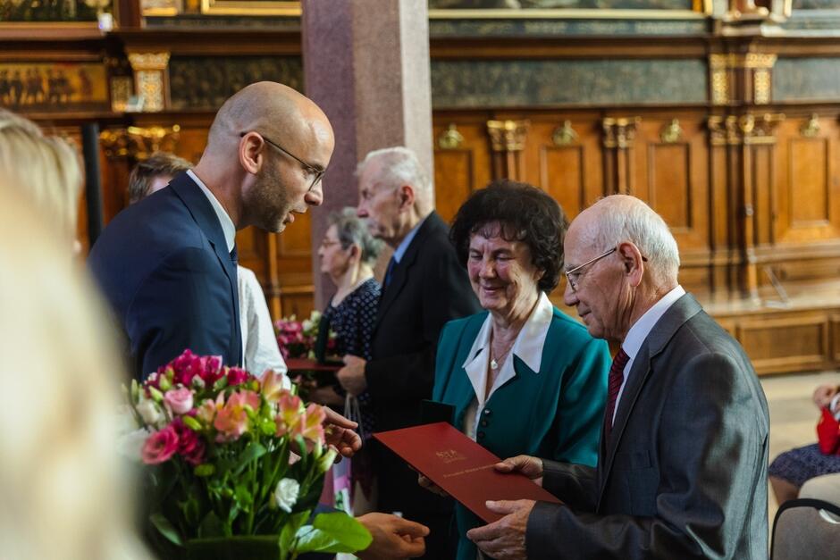 starsza para przyjmuje z rąk wysokiego mężczyzny dyplom w bordowej okładce