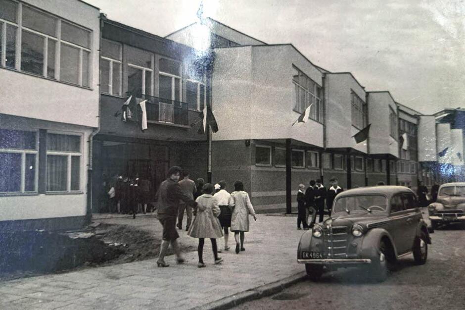 Czarno-białe zdjęcie, na którym widać budynek szkoły, kilka osób i samochód