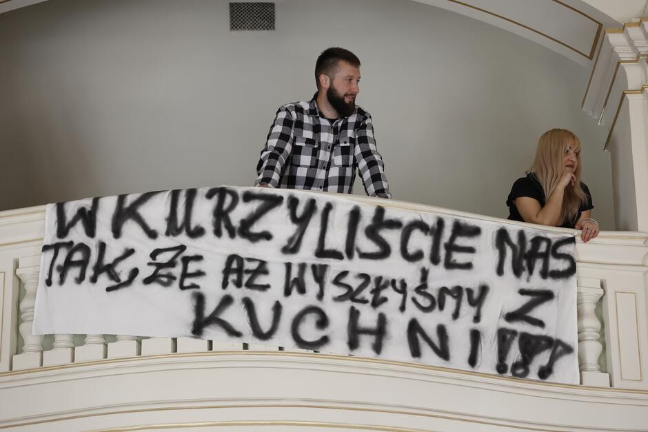Dwie osoby - kobieta i mężczyzna - na czymś w rodzaju balkonu pod sufitem sali plenarnej Rady Miasta Gdańska. Wywiesili transparent zrobiony z białego prześcieradła, czarne litery tworzą hasło: Wkurzyliście nas tak, że aż wyszliśmy z kuchni!!!