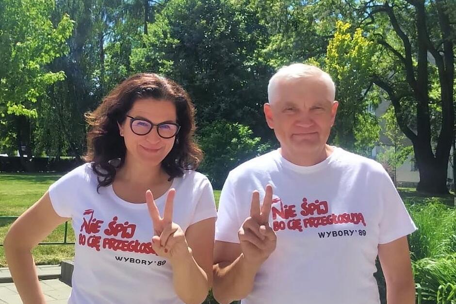 Kobieta w średnim wieku po lewej i siwy starszy mężczyzna po prawej. Pozują do zdjęcia w białych koszulkach z czerwonym napisem NIE ŚPIJ, BO CIĘ PRZEGŁOSUJĄ