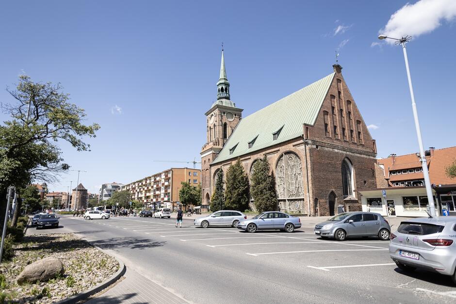 na zdjęciu fragment ulicy i kościół ceglany, w tle fragmenty niewysokich budynków mieszkalnych