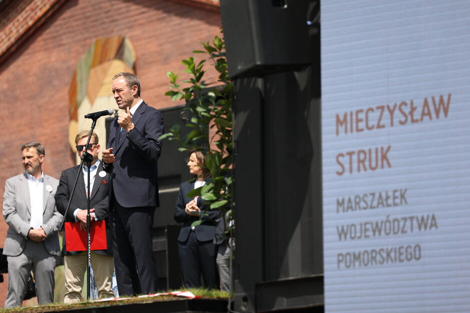 wysoki mężczyzna w garniturze mówi do mikrofonu na scenie, obok plansza z podpisem Mieczysław Struk