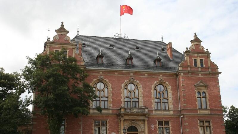 Widok zabytkowego reprezentacyjnego budynku z czerwonej cegły, ze spadzistym dachem. Budynek ma dwa piętra. Na dachu znajduje się maszt z flagą Gdańska 
