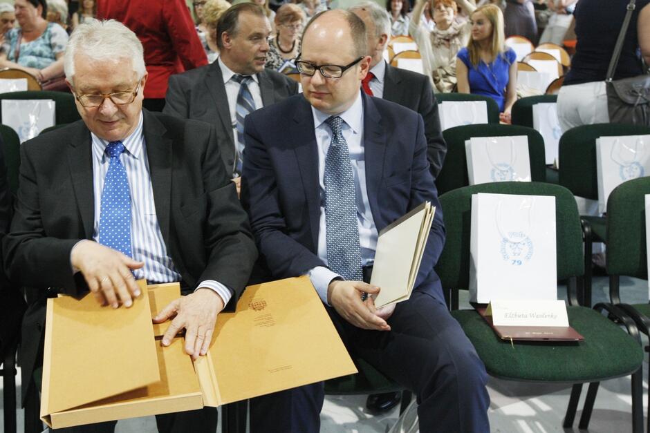 Dwóch mężczyzn siedzących na krzesłach, z których jeden ogląda pamiątkową teczkę z dokumentami 