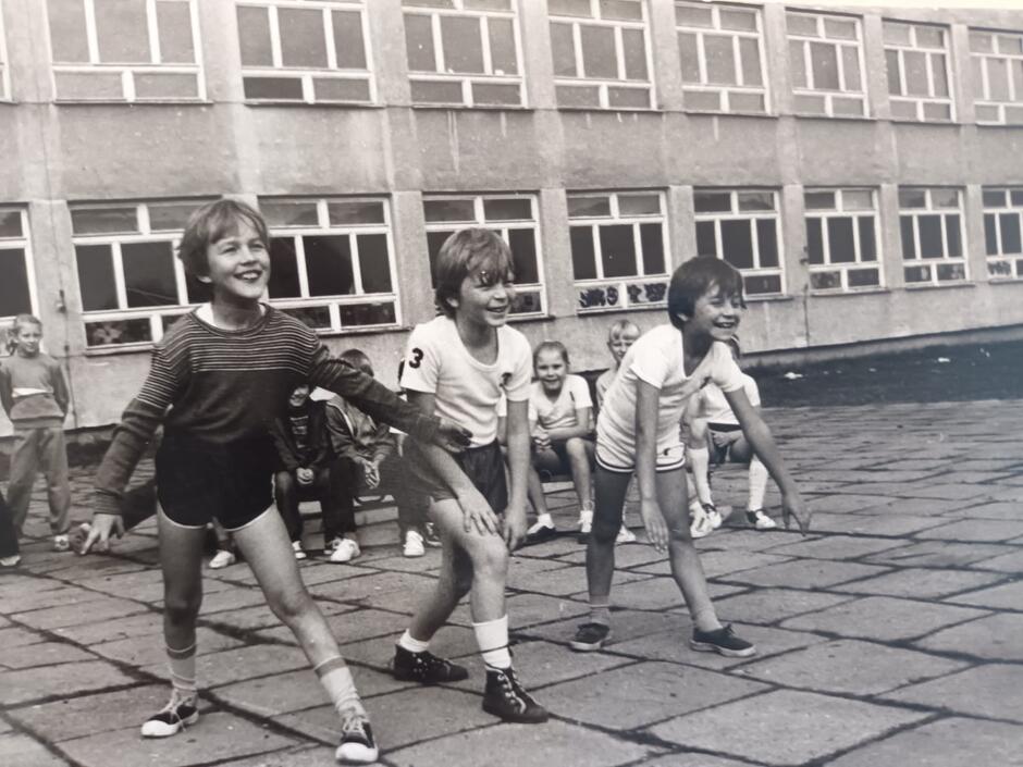 Czarno-białe zdjęcie przedstawiające kilka małych dzieci wykonujących ćwiczenia gimnastyczne przed szkołą