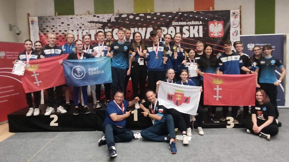 Kilkanaście osób na sali gimnastycznej siedzi lub stoi z medalami na szyjach, niektórzy trzymają w dłoniach flagi Gdańska