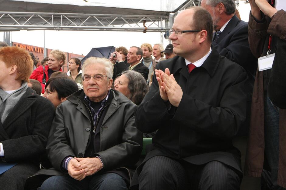 W pierwszym planie widzimy dwie siedzące osoby - centralnie: starszy, siwy mężczyzna w czarnych okularach, ubrany w brązową kurtkę. To Frank Meisler. Na prawo od niego duży mężczyzna w średnim wieku i jasnych okularach, ubrany w ciemny płaszcz - to prezydent Paweł Adamowicz. W tle widoczni są liczni inni uczestnicy uroczystości