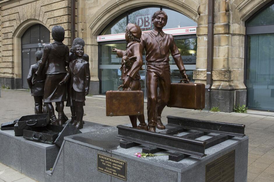Kilka figur dzieci naturalnej wielkości, które są rzeźbami, wykonanymi w brązie. Sfotografowano je na tle jednego z budynków dworcowych w Hamburgu 