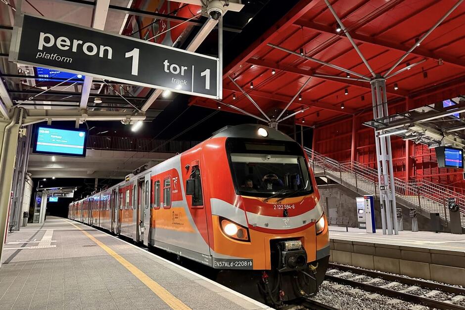 Pomarańczowy pociąg elektryczny stoi na peronie dworca. Jest noc
