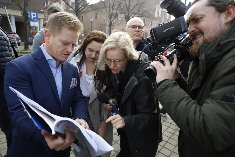 Mężczyzna w średnim wieku trzyma grubą zszywkę dokumentów, które prezentuje dziennikarzom. Mężczyzna stoi po lewej, dziennikarze są po prawej - kilka osób, mężczyzna stojący skrajnie po prawej nagrywa tę sytuację kamerą 