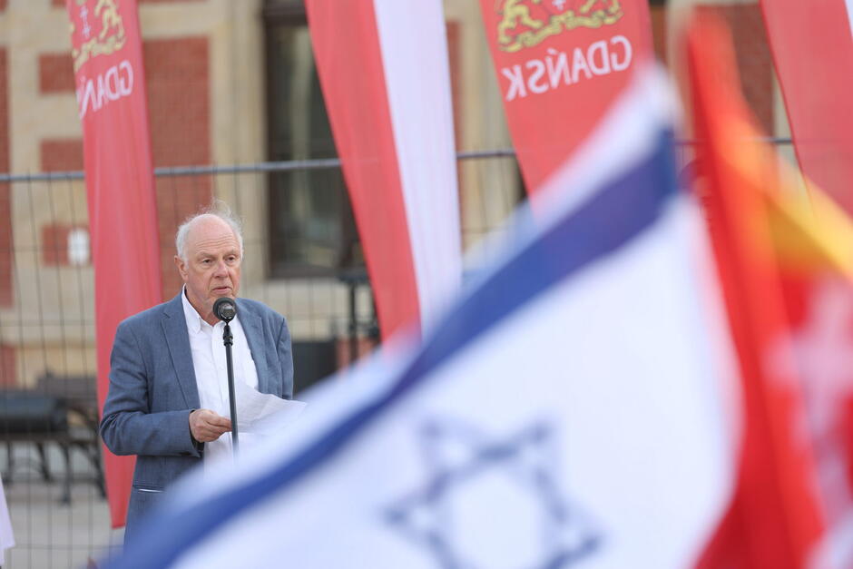 Siwy mężczyzna przed mikrofonem, przed nim flaga żydowska