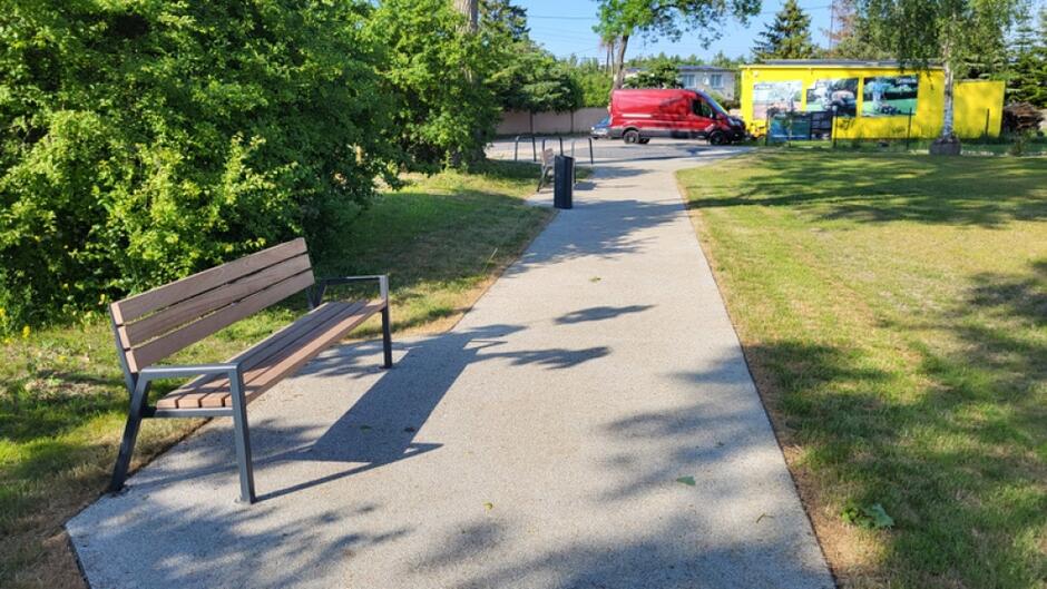 na zdjęciu fragment ścieżki dla pieszych przy niej stoi ławka po lewej, po prawej fragment zielonego trawnika, w tle samochód i mały budynek