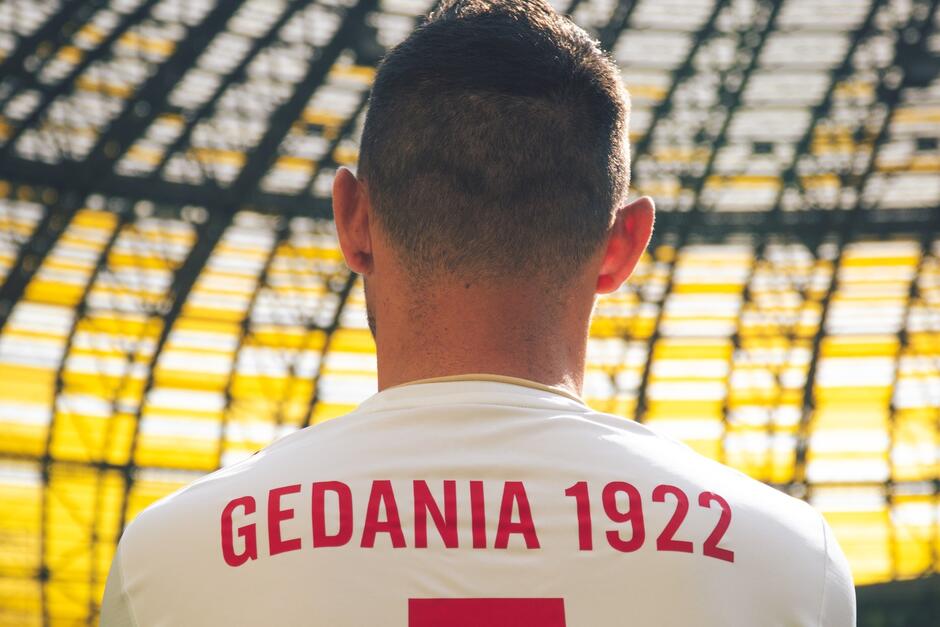 Zdjęcie przedstawia plecy i tył głowy piłkarza, które widać na tle korony stadionu Polsat Plus Arena Gdańsk. Koszulka zawodnika jest biała, w górnej części pleców znajduje się czerwony napis Gedania 1922, niżej widzimy górny fragment czerwonego numeru 5 