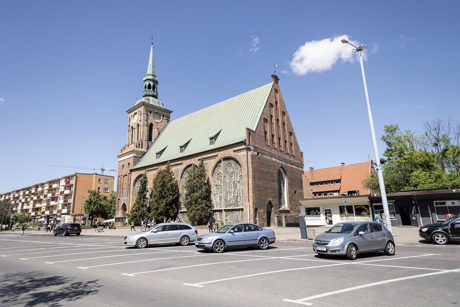 na zdjęciu fragment ulicy i kilka zaparkowanych samochodów w wyznaczonych miejscach na środku jezdni, w tle kościół