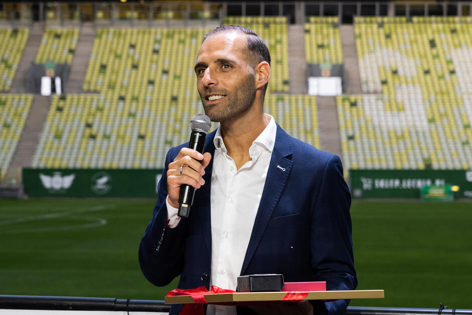 Uśmiechnięty mężczyzna w garniturze przed mikrofonem, w tle trybuny stadionu