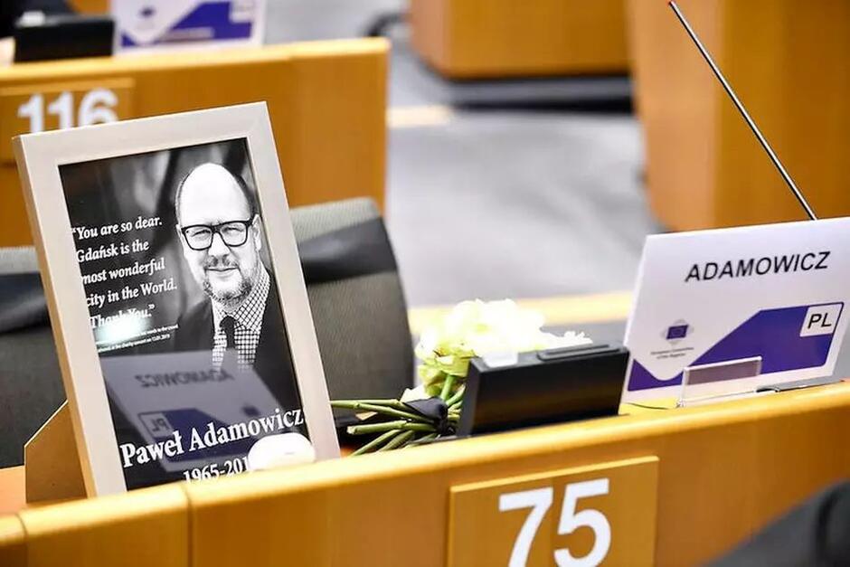 sala plenarna, miejsce za pulpitem ze zdjęciem uśmiechniętego mężczyzny w średnim wieku, obok tabliczka z napisem Adamowicz  i kwiaty