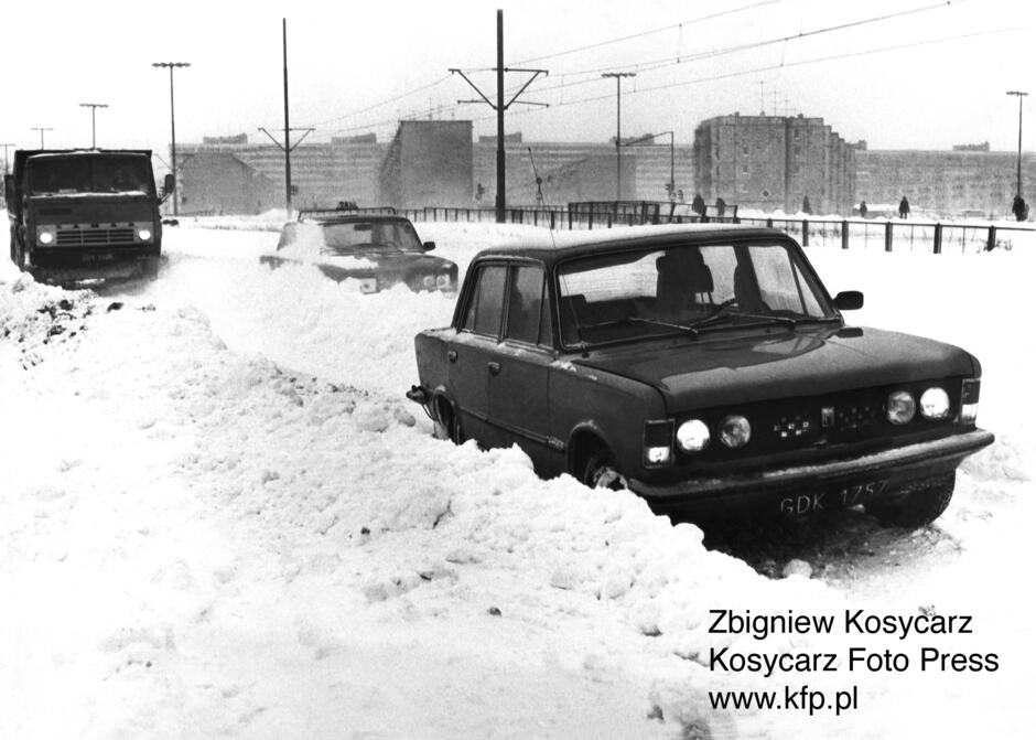 czarno białe-zimowe zdjęcie, w ogromnym śniegu utknął samochód, w oddali bloki