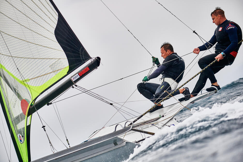 Dwóch żeglarzy na łódce (po prawej), ciągną linki żagli (po lewej)