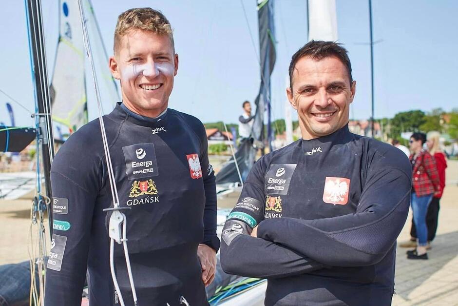Dwóch mężczyzn stoi obok siebie, w tle żaglówki