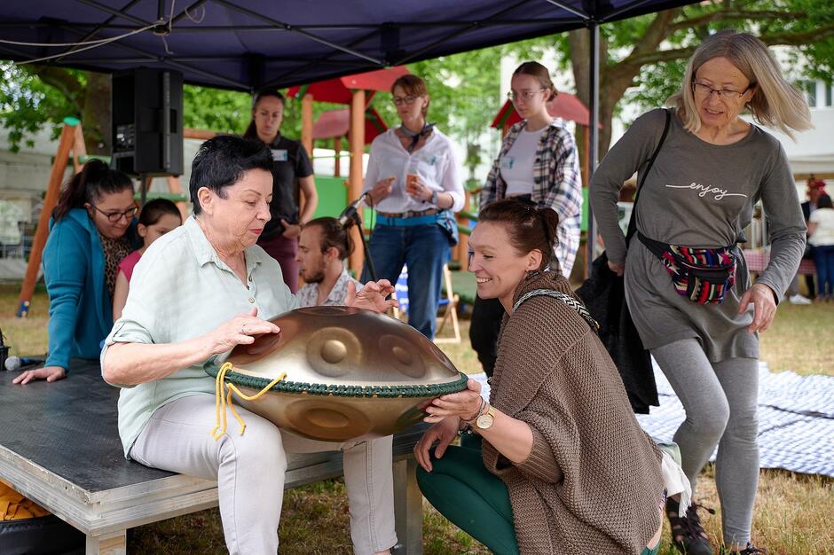 ludzie siedzą na podeście na podwórzu, jedna kobieta trzyma misy do grania