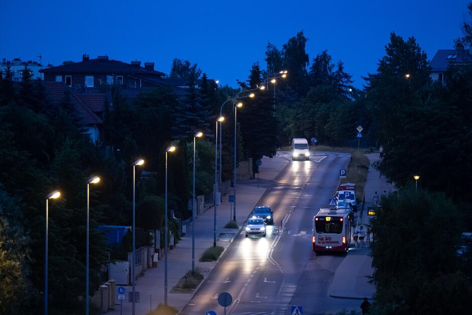 na zdjęciu ulica po zmierzchu, widać świecące latarnie, fragment jezdni i chodnika, jadące samochody i autobus miejski