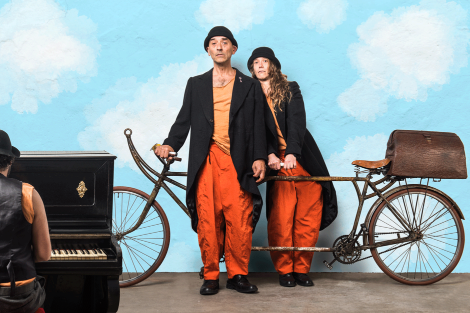 Kobieta i mężczyzna na zdjęciu są ubrani we fraki i cylindry, stoją opierając się o stary rower