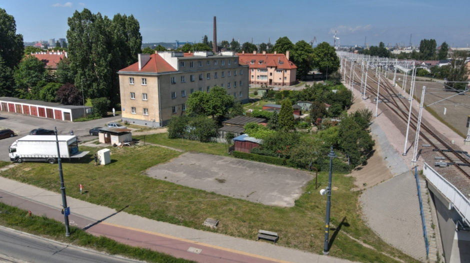 na zdjęciu z drona widać zielony teren, zniszczone boisko betonowe, po prawej na nasypie leżą tory kolejowe, a po lewej widać budynki mieszkalne i zaparkowane obok samochody