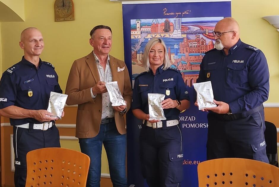 Od lewej mężczyzna w mundurze, w średnim wieku, łysy obok mężczyzna w średnim wieku z krótkimi włosami w marynarce, obok młoda kobieta w mundurze, obok niej postawny mężczyna w mundurze