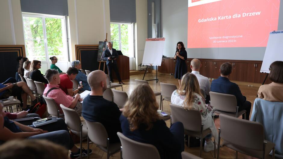 Kobieta w okularach przemawiająca na stojąco do mikrofonu do kilkunastu osób siedzących na krzesłach w sali