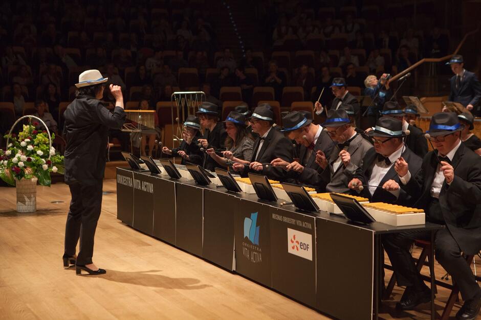 kobieta dyryguje, przed nią siedzą muzycy