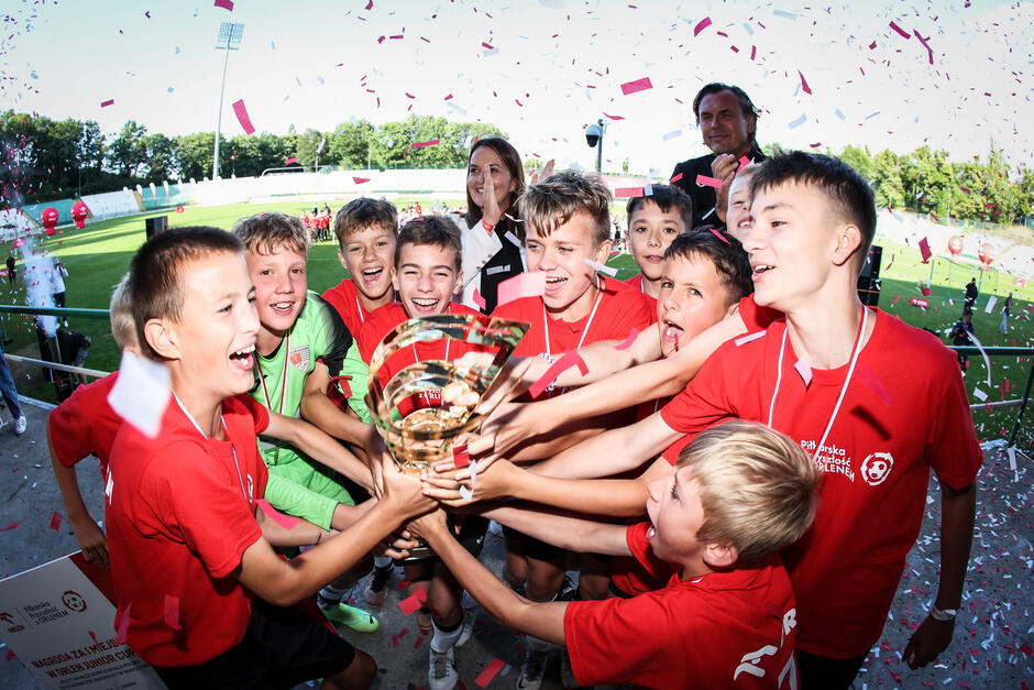Chłopcy w czerwonych koszulkach stoją w kręgu, cieszą się, trzymają duży puchar