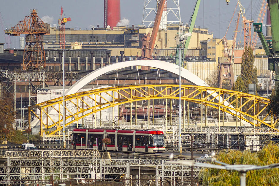 Zdjęcie przedstawia panoramę miasta - na pierwszym planie torowisko z jadącym tramwajem. Dalej widać żółty stalowy wiadukt nad torowiskiem kolejowym i za nim jasny wiadukt ul. Nowa Wałowa. Dalej budynki i kominy przemysłowe oraz dźwigi stoczniowe. Kolejne perspektywy nakładają się na siebie, dając efekt rosnącego obrazu 