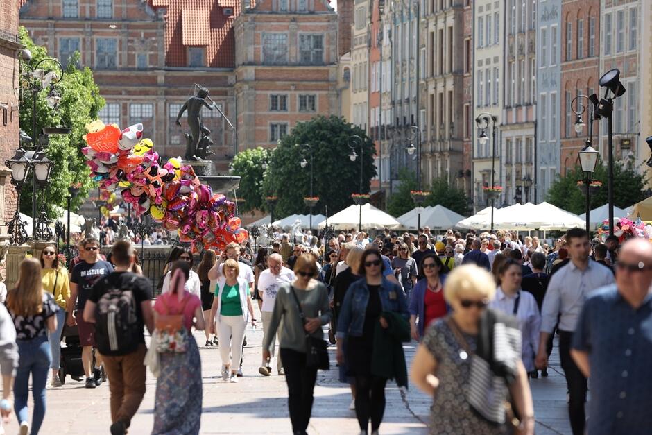 Na pierwszym planie wiele osob w letnich ubraniach w różnym wieku spaceruje po ulicy, z prawej i lewej stronie stare kamienice