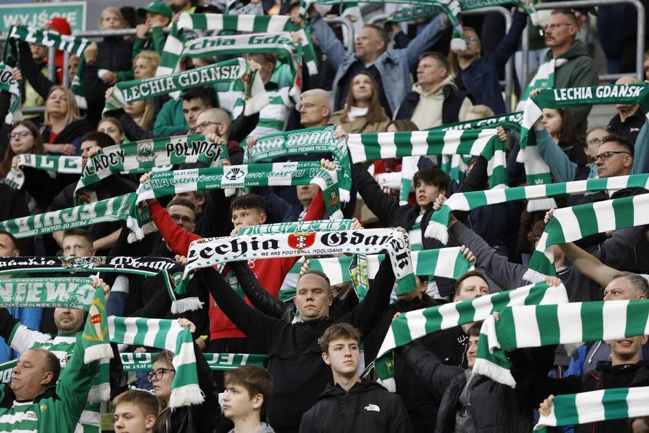 Zdjęcie przedstawia tłum kibiców na trybunie stadionu, którzy stoją unosząc w górę biało-zielone szaliki z napisami Lechia Gdańsk. Szaliki trzymane są przez wszystkich oburącz