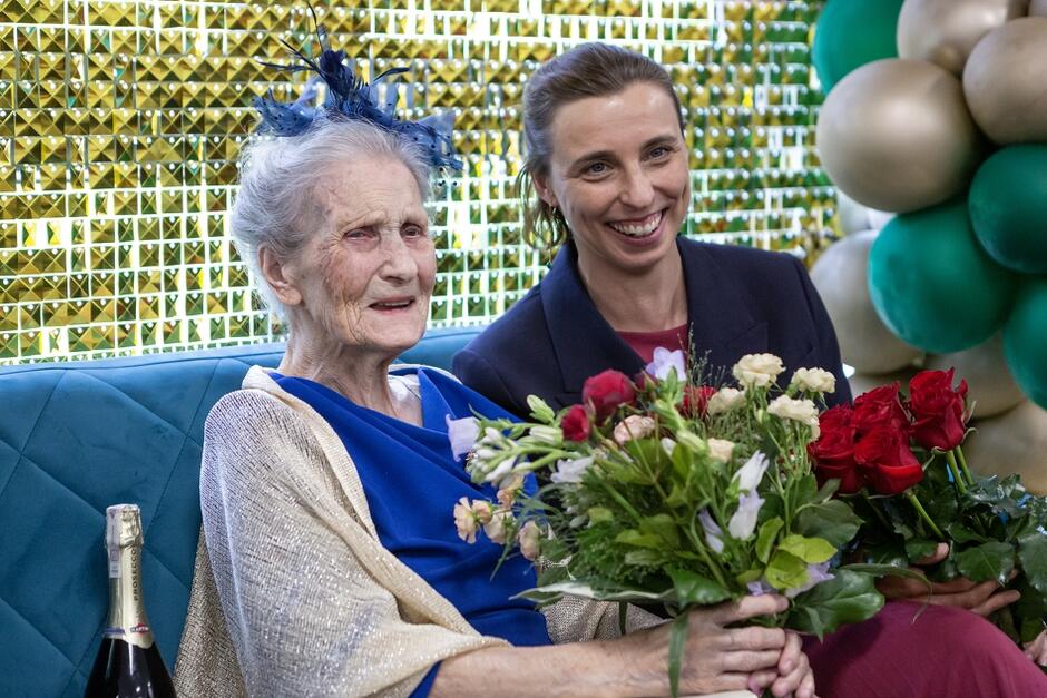 starsza elegancko ubrana kobieta z kwiatami, obok niej młodsza kobieta