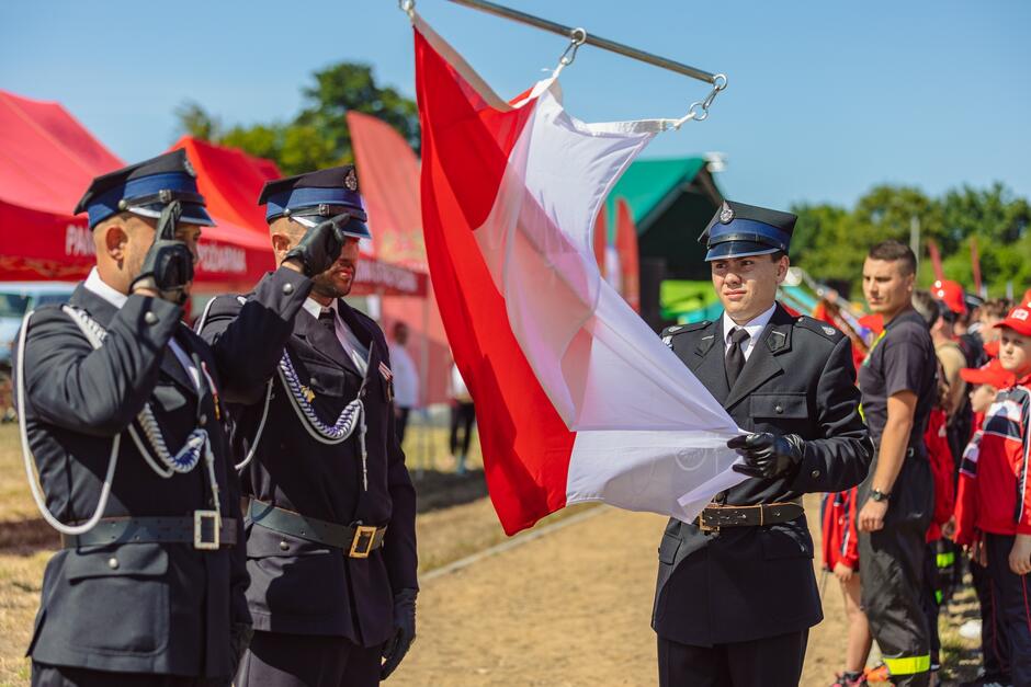 trzech strażaków w mundurach galowych, dwóch salutuje trzymanej przez trzeciego fladze Polski