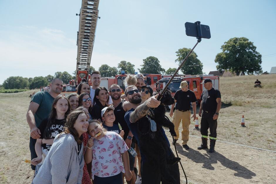 kilkanaście osób pozuje, jedna z nich trzyma selfiestick, na tle wozów strażackich