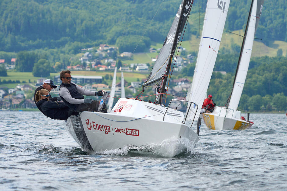 Troje żeglarzy wychylonych za łódką z żaglem. W tle inne łodzie