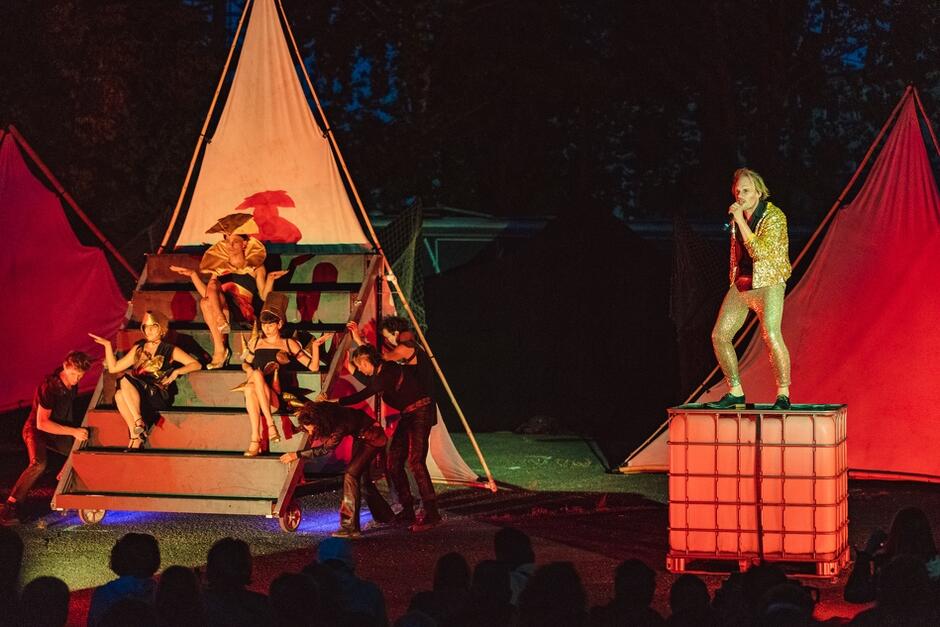 na platformie w kształcie piramidy siedzą aktorzy, obok na dużej skrzynce śpiewa aktor w złotym garniturze