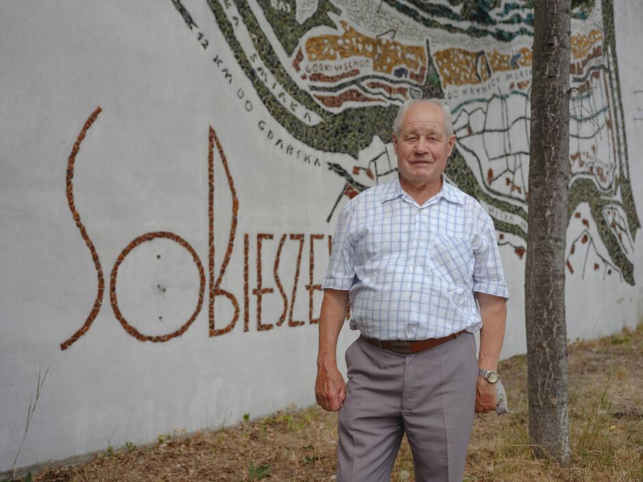 starszy pan stoi na tle napisu na ścianie Sobieszewo