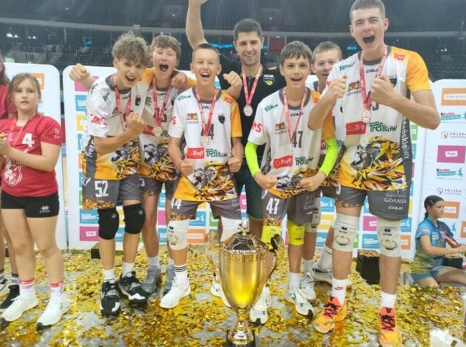 Kilku chłopców w strojach sportowych stoi obok siebie, cieszą się, na szyjach mają medale. Przed nimi stoi puchar