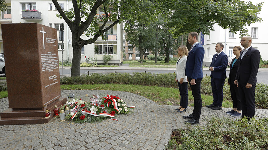Kilka osób stojących w postawie na baczność przed pomnikiem, pod którym leżą kwiaty