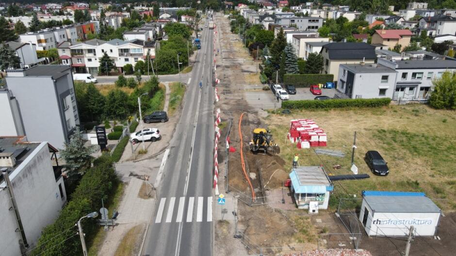 zdjęcie z drona, widać asfaltową ulicę, po prawej stronie na poboczu prowadzone są prace ziemne, w tle po obu stronach widać niewysoką zabudowę mieszkaniową