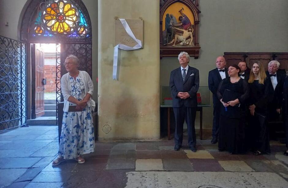 grupa osób w kościele w strojach odświętnych, na filarze przesłonięta wstęgą tablica