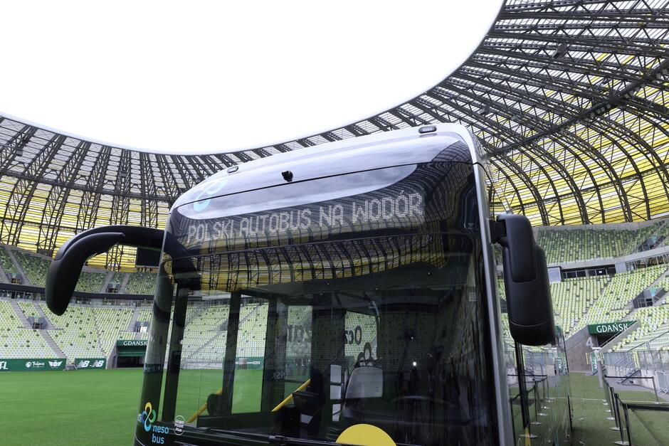 na zdjęciu fragment przedniej części ciemnego autobusu, w tle wnętrze gdańskiego stadionu